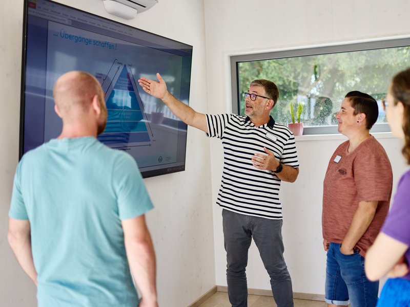 Menschengruppe steht vor Whiteboard (unscharf), jemand erklärt etwas