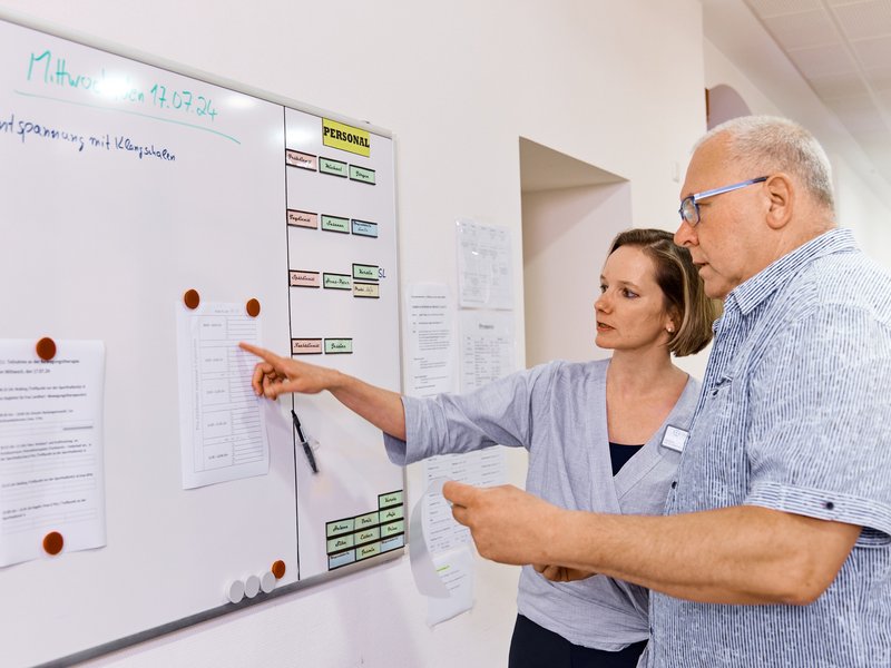 Ärztin steht mit Patient vor einer Tafel, auf der ein Therapieplan aushängt