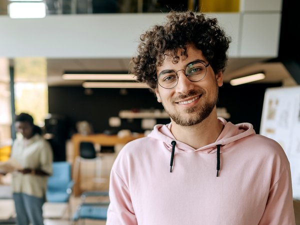 Junger Mann lächelt in die Kamera, hinter ihm unscharf eine kleine Personengruppe (Symbolbild)