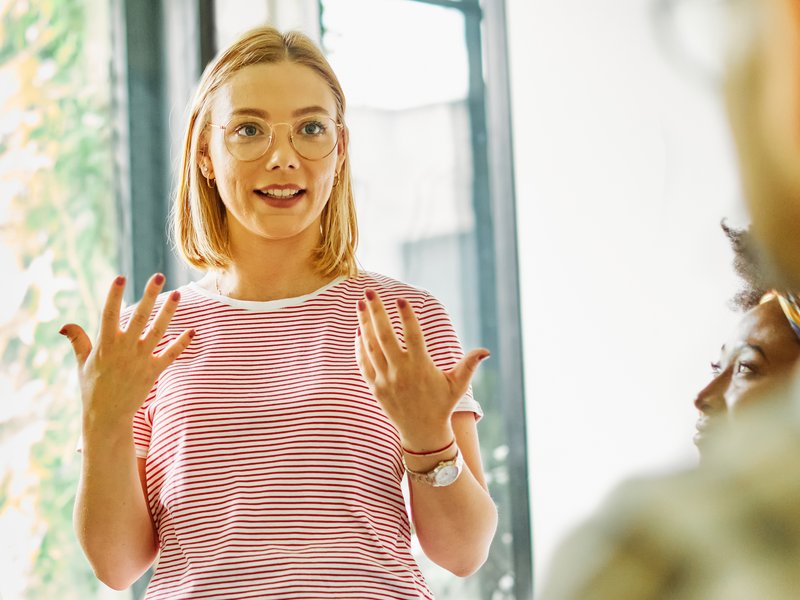 Junge Frau gestikuliert und steht in einer Gesprächsrunde, deren Teilnehmende man nicht sieht. Stockfoto.