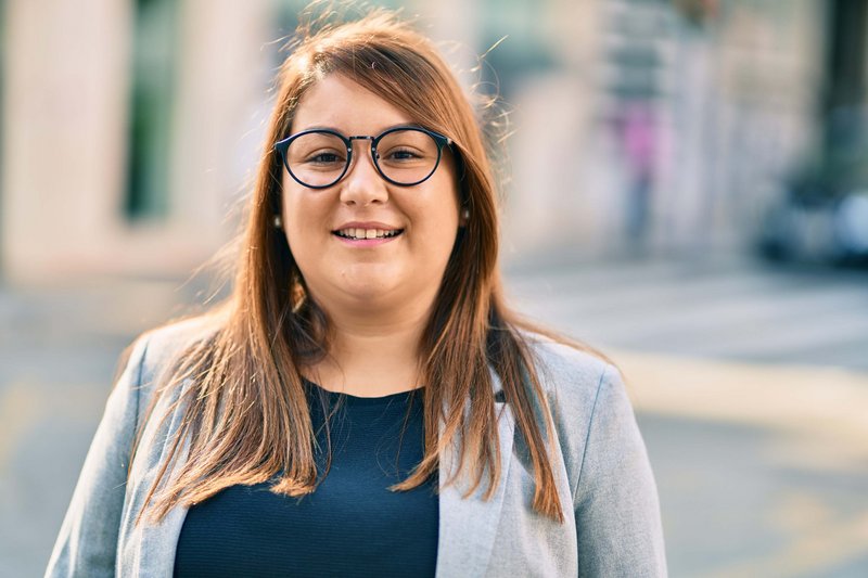 Portrait einer Studierenden mit einem blauen Oberteil und Brille vor einem unscharfen Hintergrund