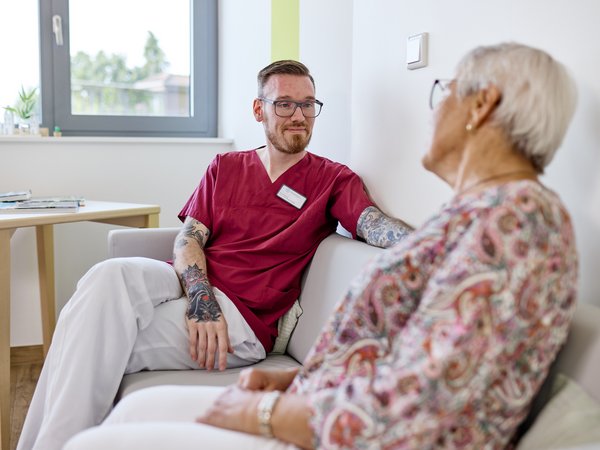 Junge Pflegekraft oder Praktikant sitzt mit älterer Dame für ein Gespräch auf einem Sofa