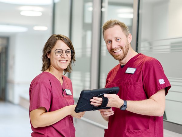 Zwei Pflegekräfte stehen im Stationsflur mit einem Tablet und lächeln in die Kamera
