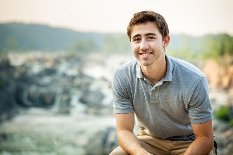 Portrait eines jungen Mannes in der Natur. Er sitzt entspannt und lächelt in die Kamera.